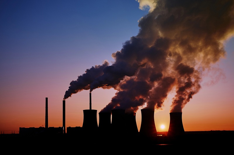 Silhouetted industrial plant emitting thick smoke against a vibrant sunset sky, depicting pollution and environmental concerns amid an economic slowdown